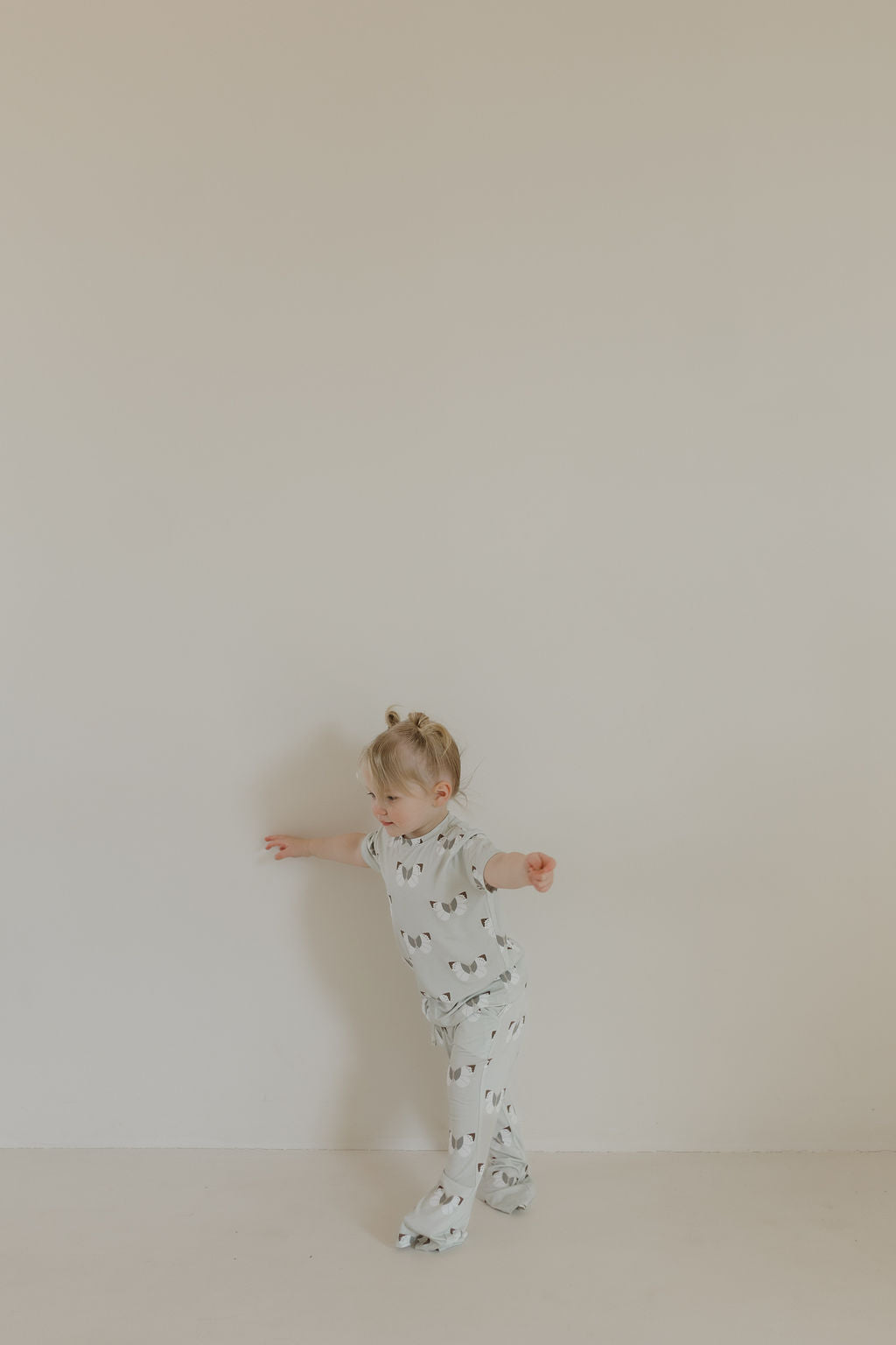 A young child with blonde hair wears a hypoallergenic Toddler Short Sleeve Bamboo Lounge Set in the Butterflies pattern by forever french baby. Barefoot, they playfully balance against a plain white wall, their arms extended as if dancing. The bright light enhances the minimalistic scene.