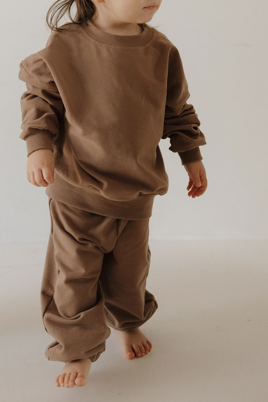 A young child, partially out of frame, stands barefoot in a minimal space wearing the forever french baby Child Sweat Set | Hot Cocoa—an oversized brown sweatshirt and matching pants with rolled-up sleeves.