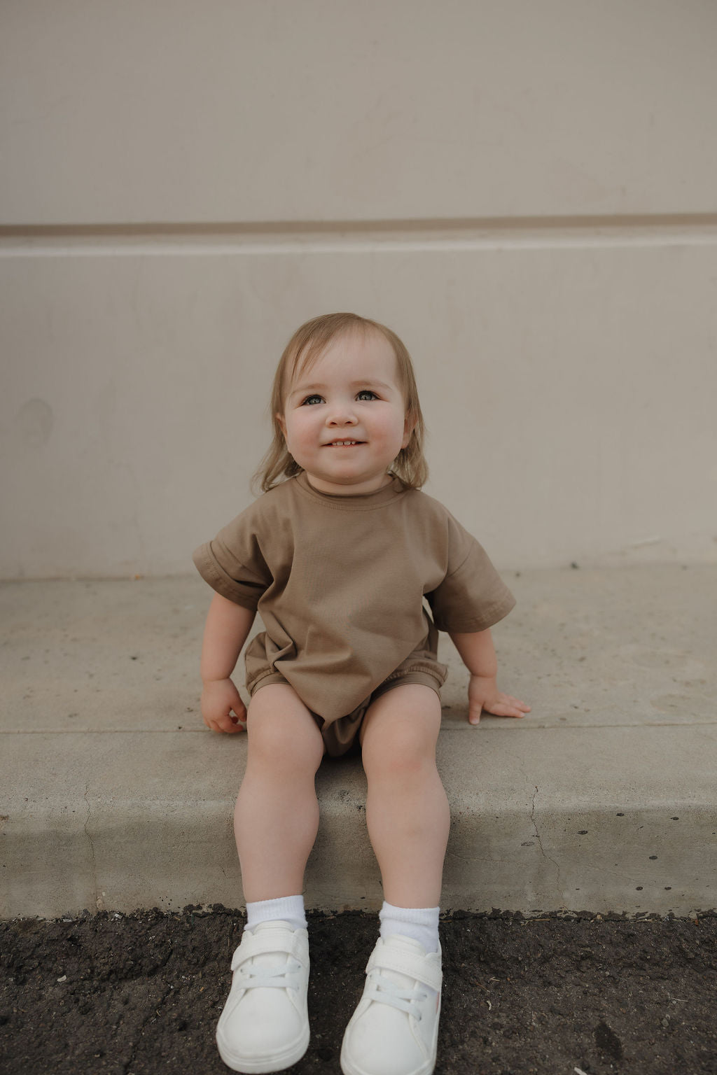 A toddler with light brown hair, wearing the forever french baby Short Sleeve Romper in Patina, white socks, and white shoes, sits smiling on a concrete curb against a light wall, looking slightly upward.