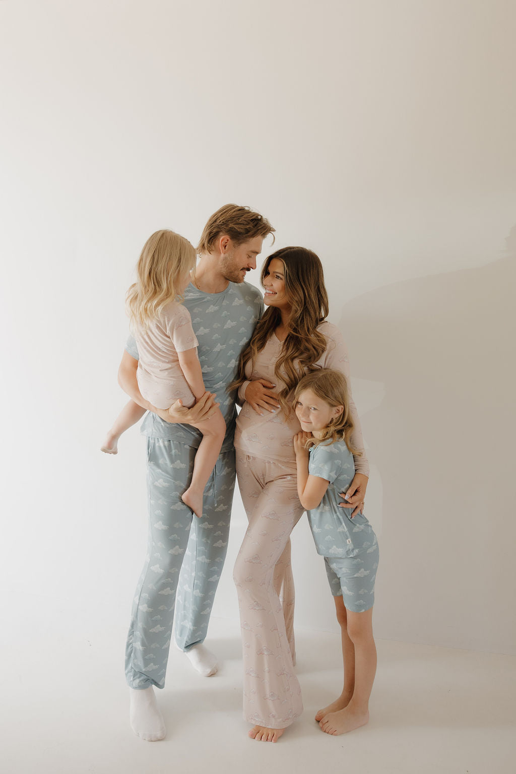 A family of four poses against a plain light background wearing Kendy 3 Bamboo Short Set | Duke pajamas—the father holds a young child, the pregnant mother stands nearby, and an older child hugs her side.