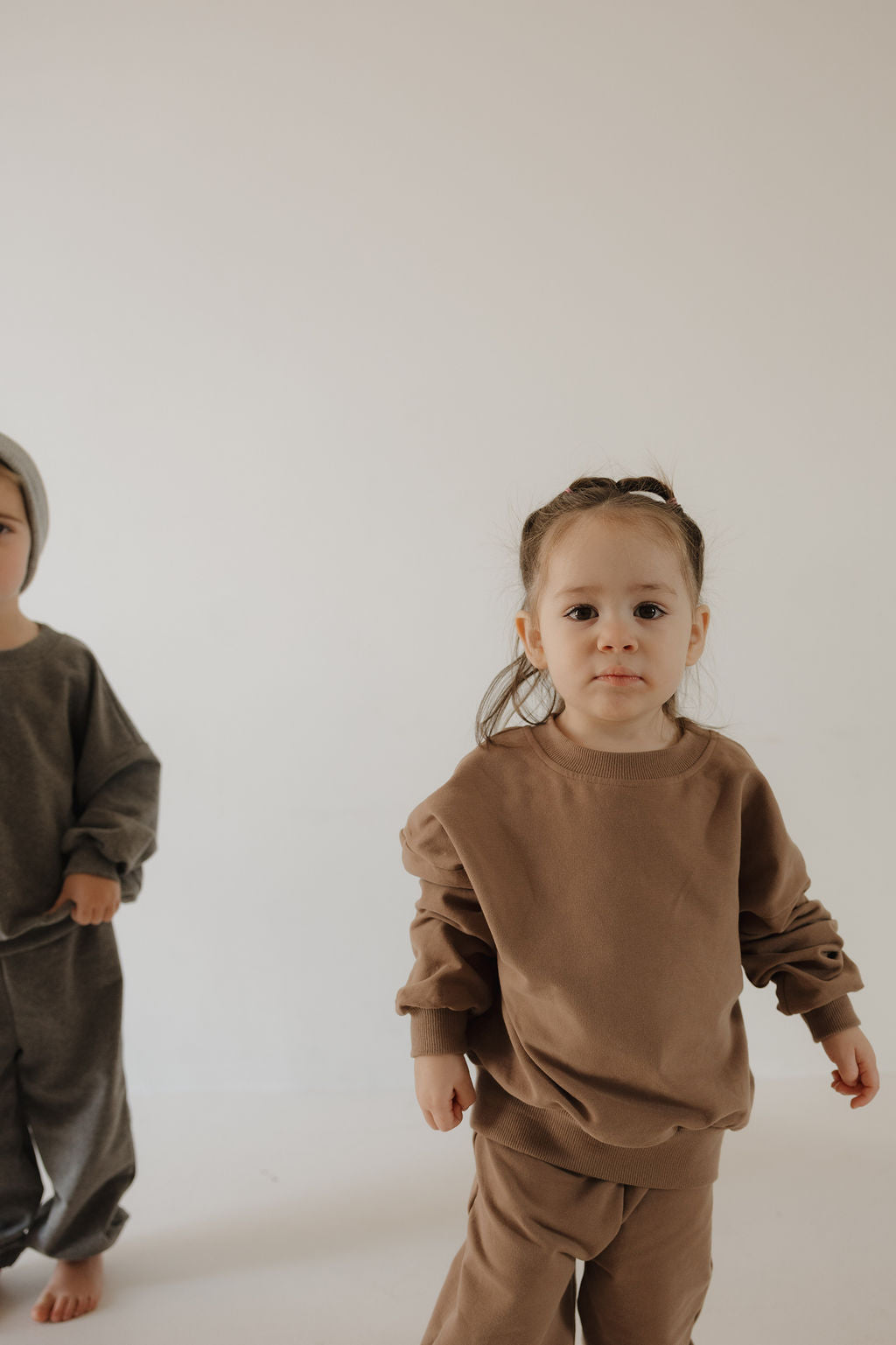 Two young children stand indoors against a plain white background. The front child wears the forever french baby Child Sweat Set | Hot Cocoa, looking at the camera, while another in grey is partially visible on the left.