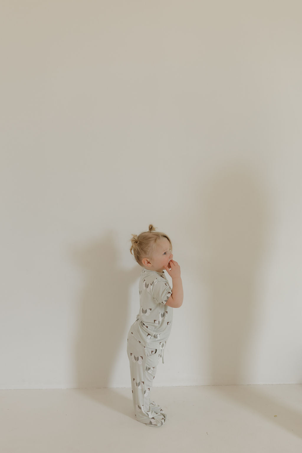 A toddler in the forever french baby Short Sleeve Bamboo Lounge Set | Butterflies stands thoughtfully against a beige wall. The hypo-allergenic, breathable bamboo pajamas suit the serene setting as her shadow gently touches the wall.