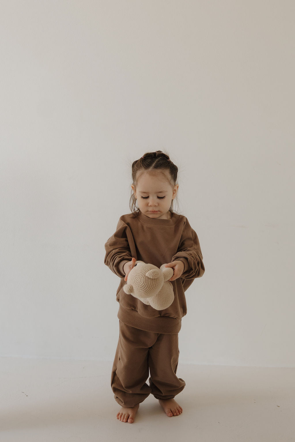 A young child wears the forever french baby Child Sweat Set in Hot Cocoa, standing barefoot on a light floor and holding a beige knitted stuffed toy against a white background.