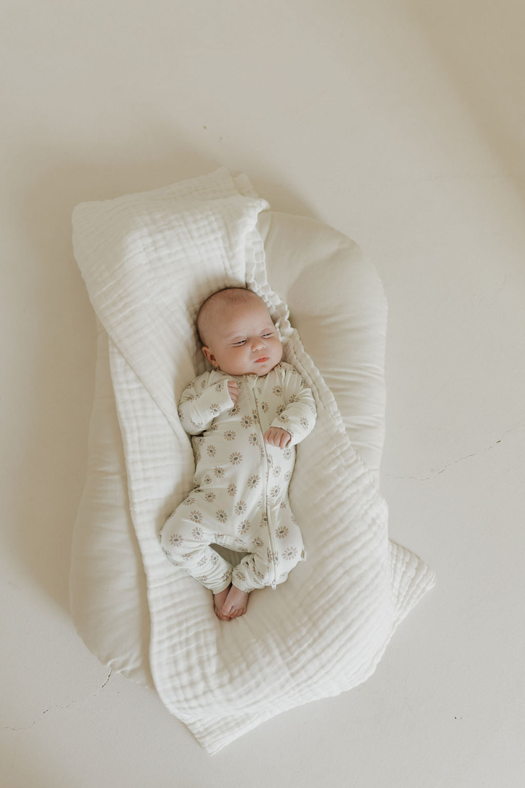 A baby drifts off peacefully on a cushioned surface, wrapped in a soft white blanket. Wearing forever french baby's hypo-allergenic Bamboo Zip Pajamas in Daisy Fields, the baby rests against a light beige background that enhances the calm and cozy atmosphere.