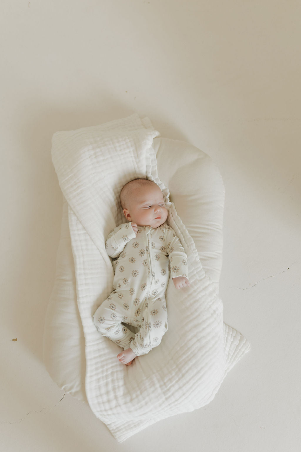 A baby wearing forever french baby's hypo-allergenic Bamboo Zip Pajamas in Daisy Fields design peacefully sleeps on a white, cushioned blanket in a serene, minimalistic setting.