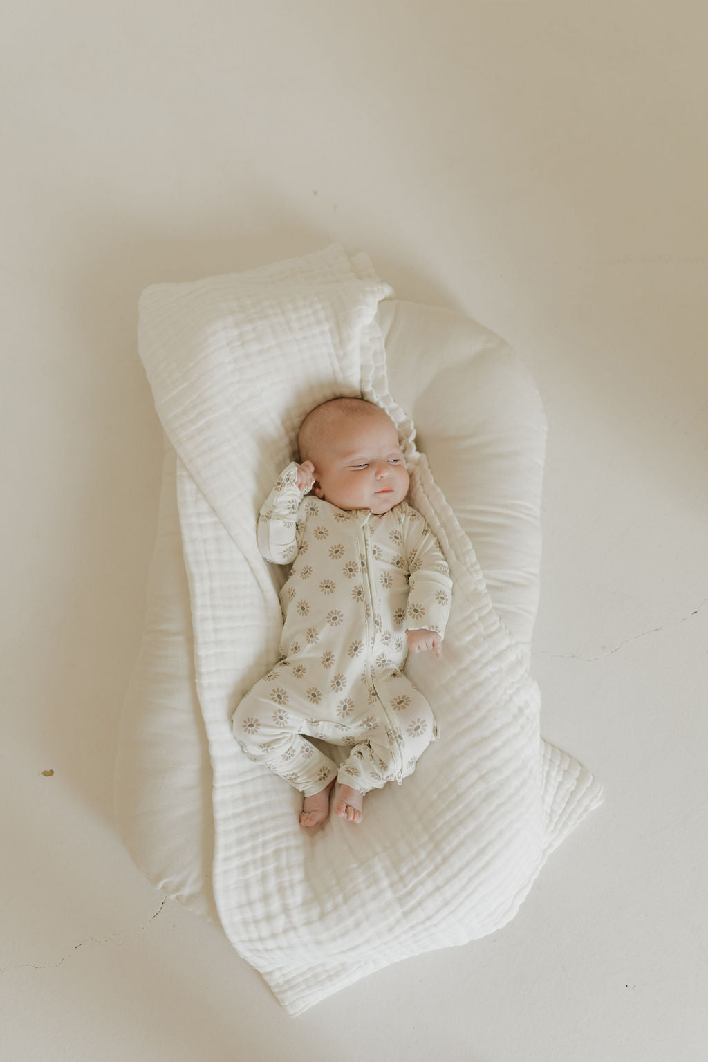 A newborn lies on a soft cream blanket, snug in forever french baby's hypoallergenic Bamboo Zip Pajamas in Daisy Fields. The baby wears a cream onesie with small floral patterns and rests peacefully with one arm slightly raised against the neutral white background.