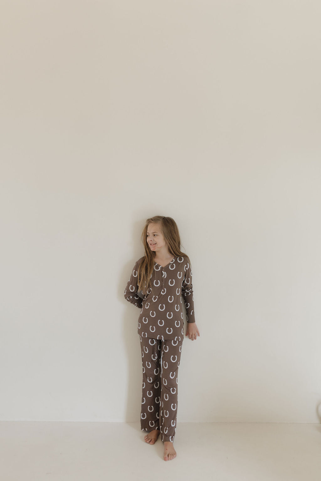 A girl with long hair, barefoot in forever french baby's Youth Flare Bamboo Pajamas—Giddy Up design, featuring brown fabric with white horseshoe patterns. Made from breathable material, she looks left while her right arm rests gently against a neutral wall.