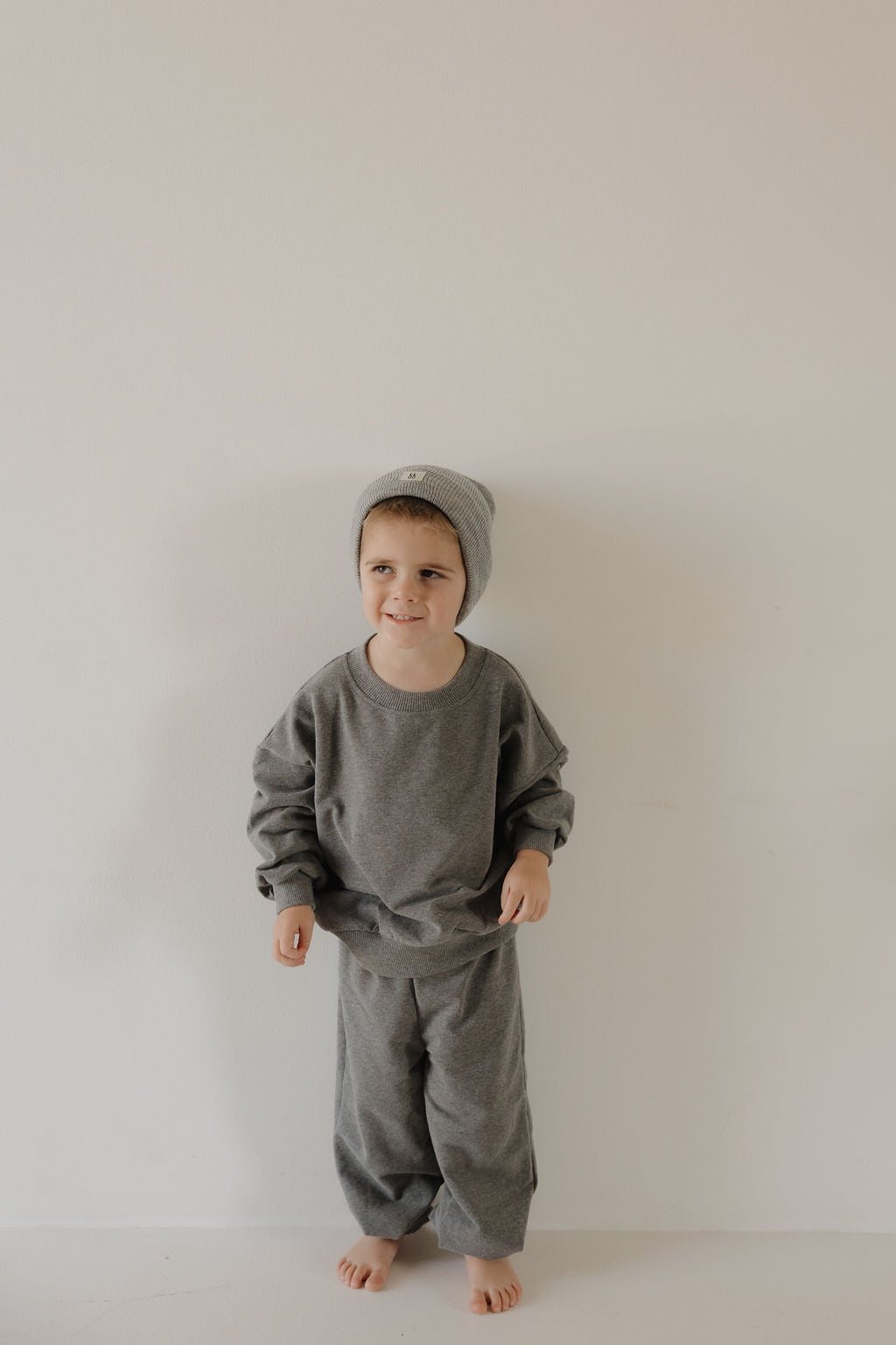 A young child stands barefoot against a light wall, smiling slightly while wearing the forever french baby Child Sweat Set in Charcoal—loose-fitting sweatshirt, matching pants, and a gray beanie.