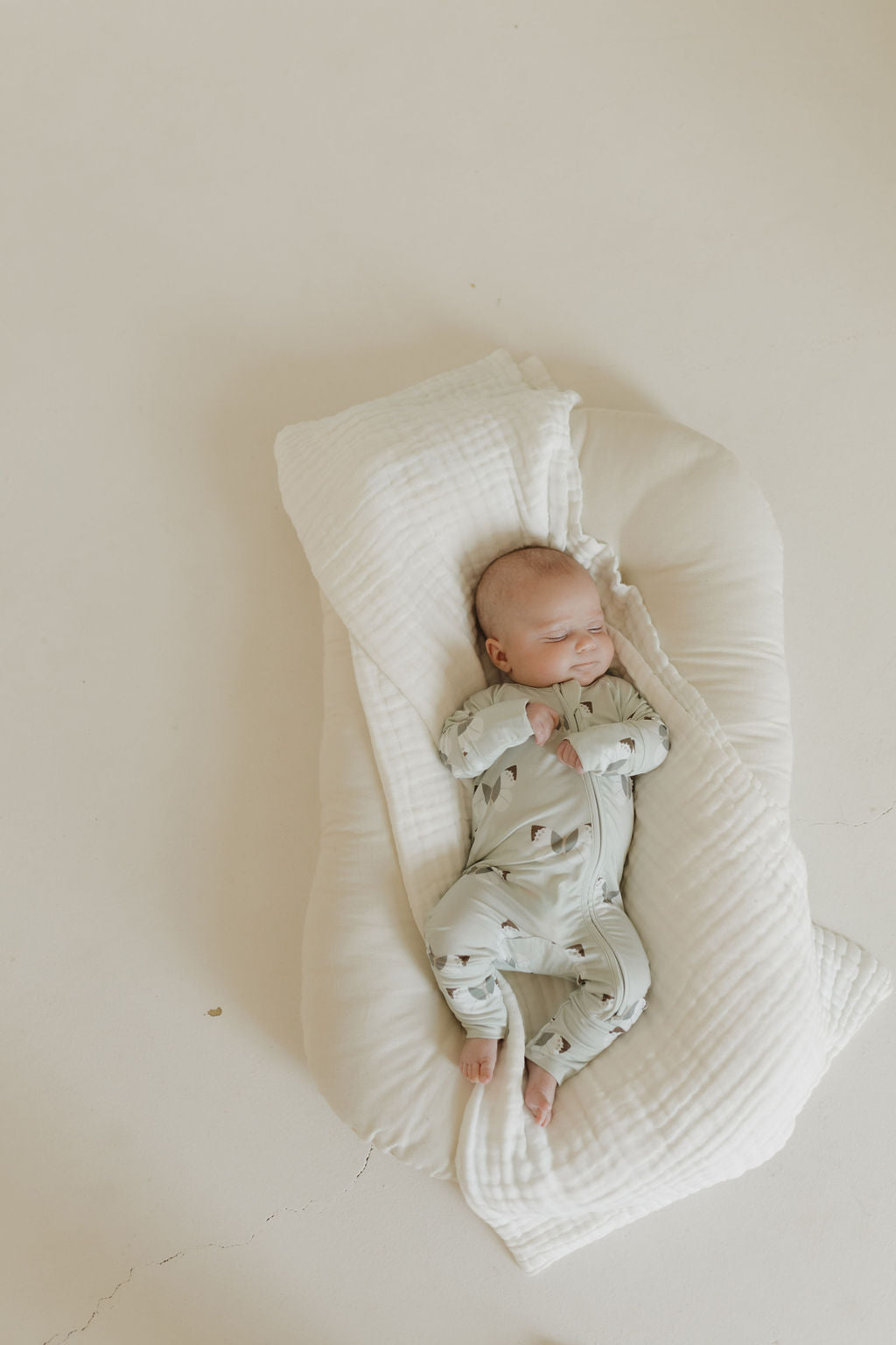 A baby sleeps peacefully on a soft, white cushion in breathable Bamboo Zip Pajamas | Butterflies by forever french baby. The light gray onesie with small patterns adds charm against the smooth, cream-colored background.