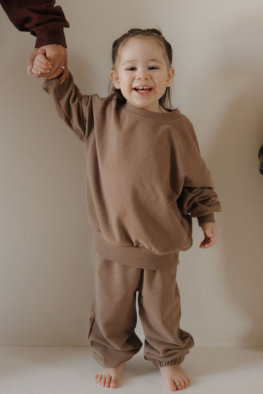 A young child beams, barefoot and holding an adult's hand, modeling the forever french baby Child Sweat Set | Hot Cocoa—versatile kids' clothing in a cozy style, perfect for playful days against any backdrop.