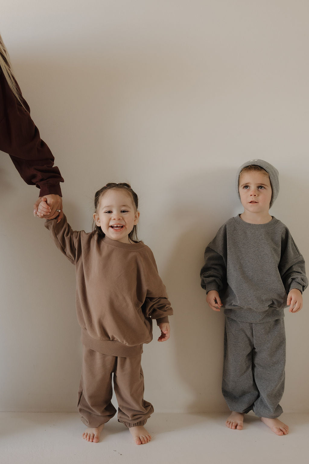 Two young children, barefoot against a light wall, wear matching forever french baby Child Sweat Set | Charcoal. The girl smiles while holding an adult’s hand; the boy pairs his set with a beanie for a coordinated family look.