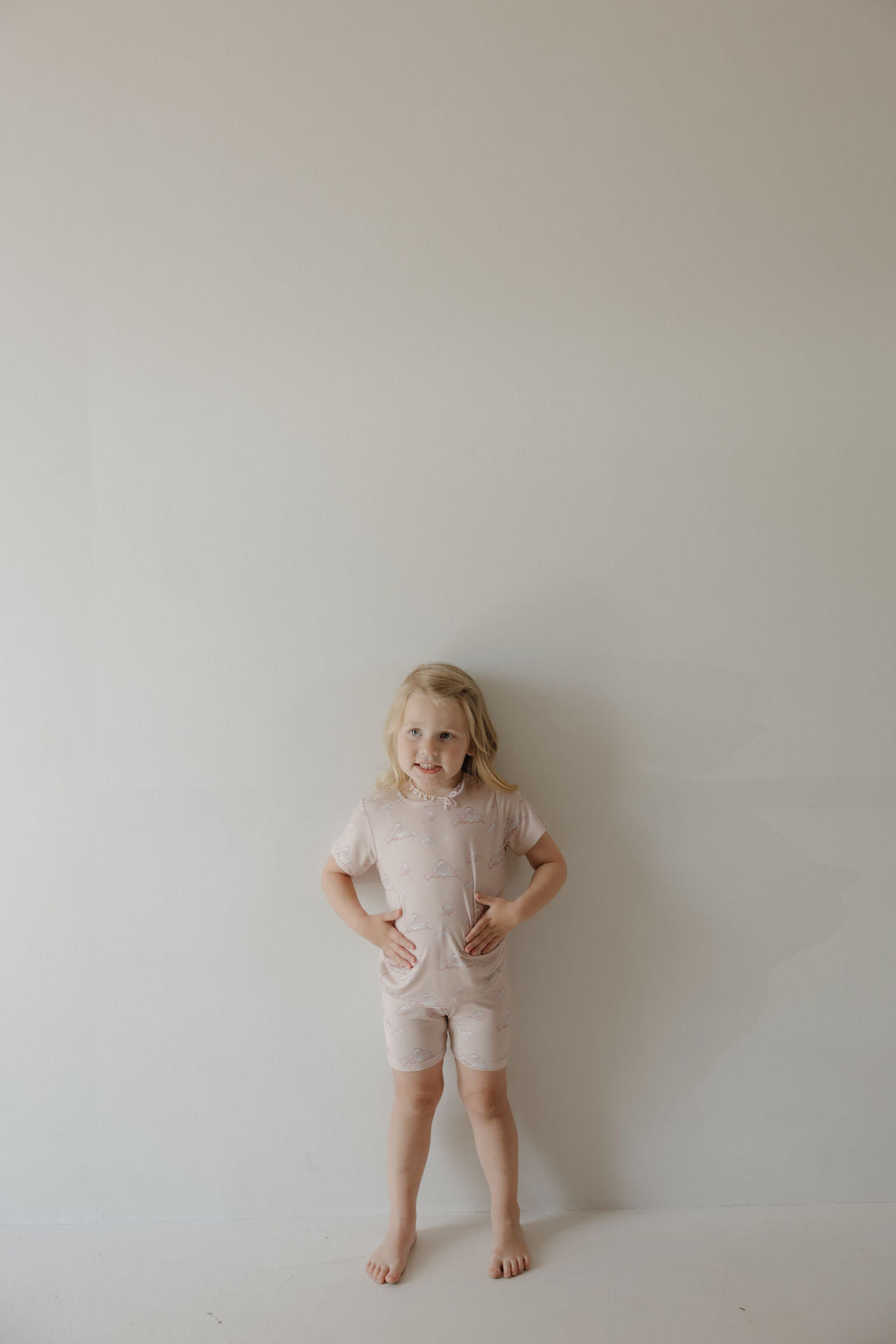 A young child with shoulder-length blonde hair stands barefoot against a light wall, smiling hands on hips while wearing the Kendy 3 Bamboo Short Set | Ruby—a breathable, light pink matching t-shirt and shorts set.