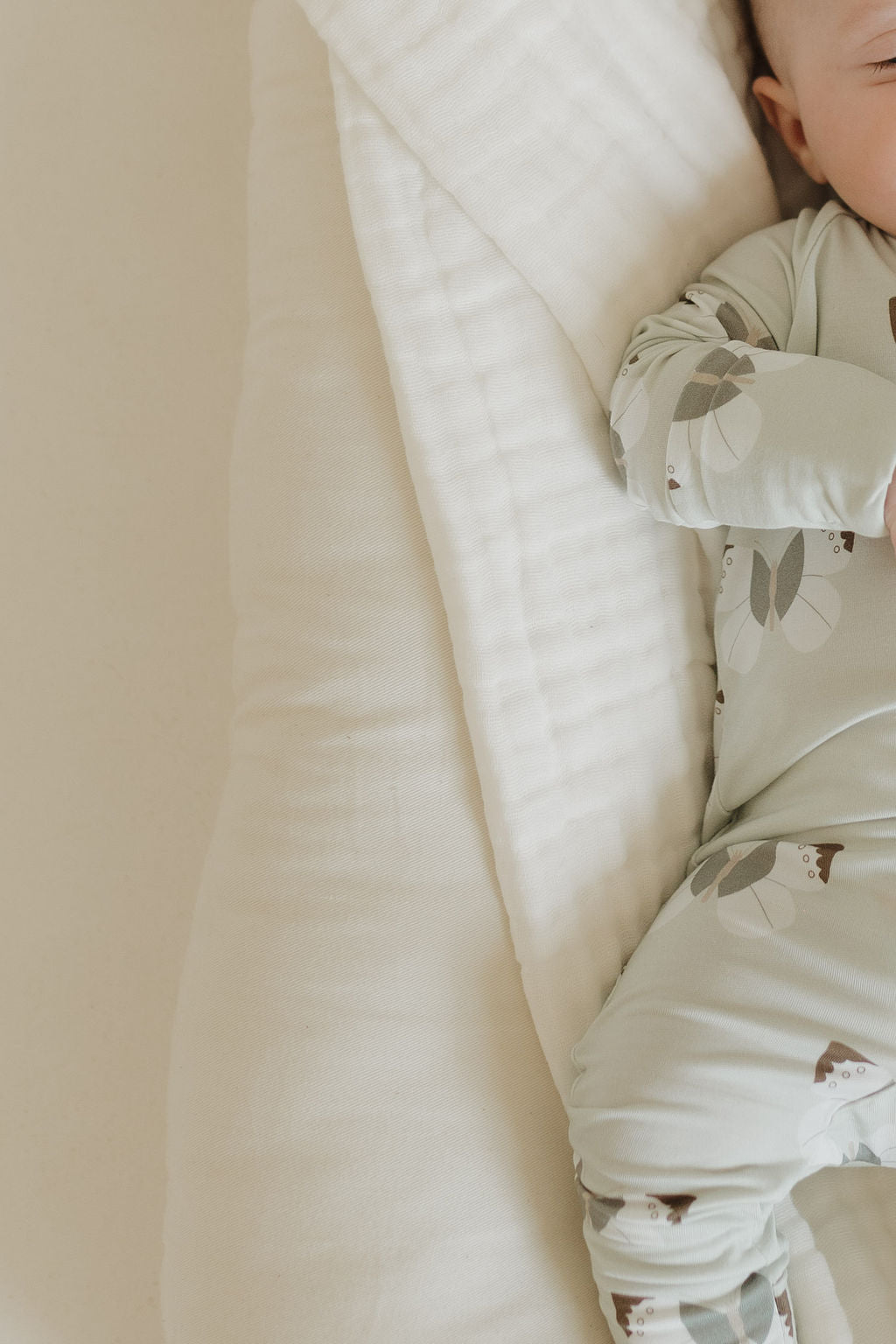 A baby in "Bamboo Zip Pajamas | Butterflies" by forever french baby rests on a soft off-white blanket. Only an arm and leg are visible, emphasizing the breathable sleepwear. The scene conveys calmness and comfort.