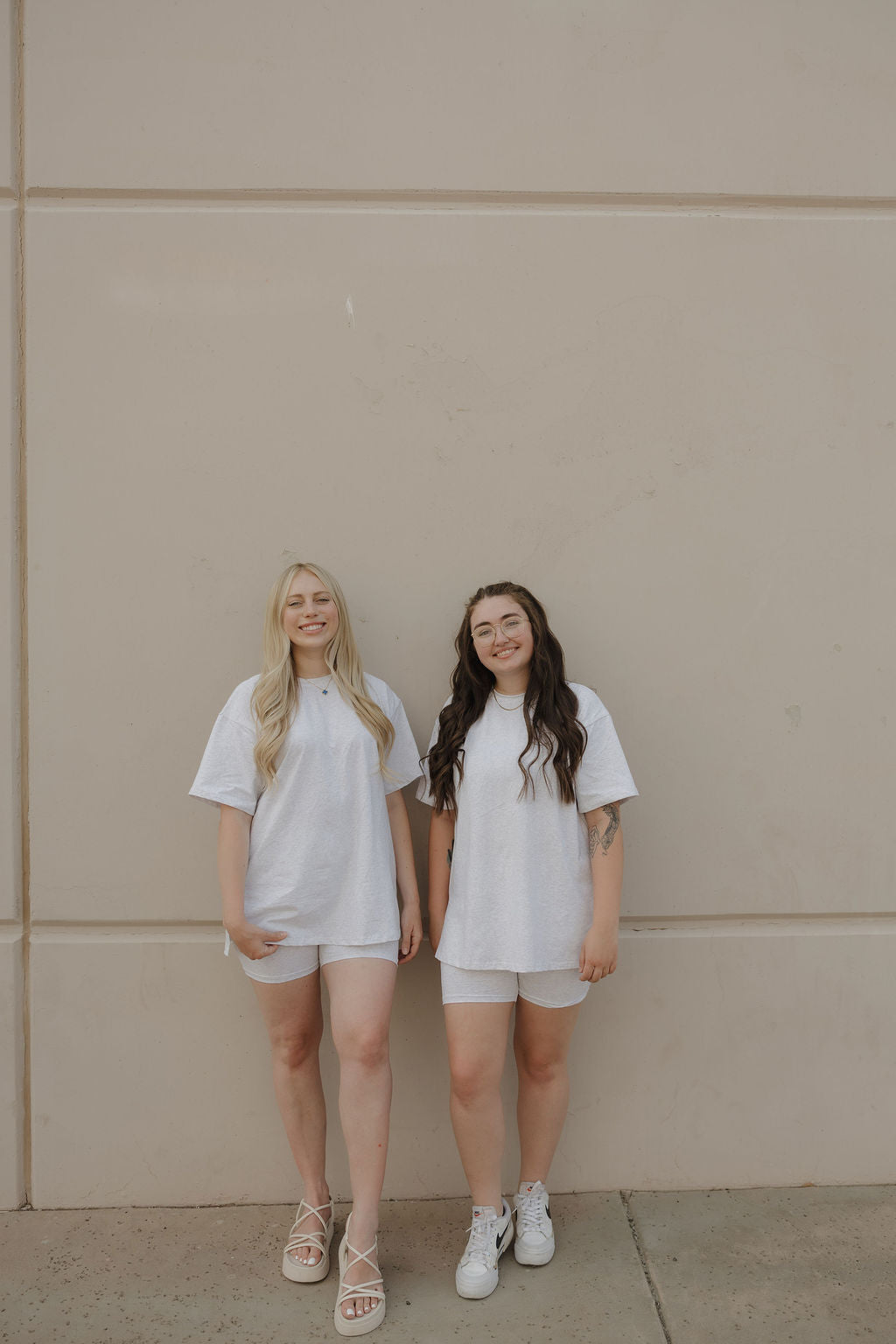 Two women stand against a beige wall wearing forever french baby’s Women's Short Set in Heather. One pairs her oversized tee with biker shorts and sandals; the other wears classic shorts with white sneakers. They smile, looking relaxed.