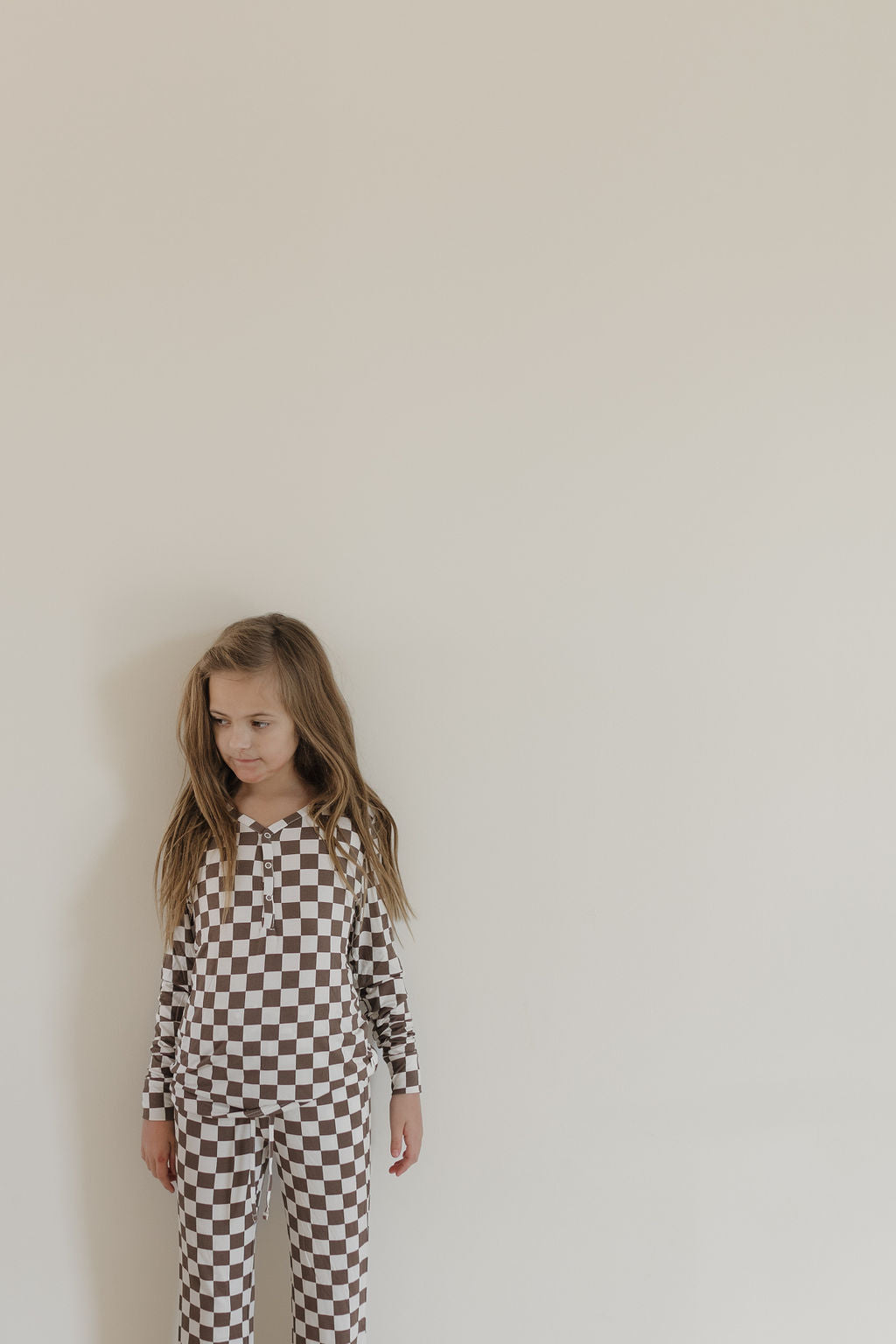 A young girl with long hair stands against an off-white wall, wearing Youth Flare Bamboo Pajamas in a brown and white checkered pattern by forever french baby. She looks to the side with a slight smile. The hypoallergenic fabric ensures comfort and style in the simple, plain backdrop.