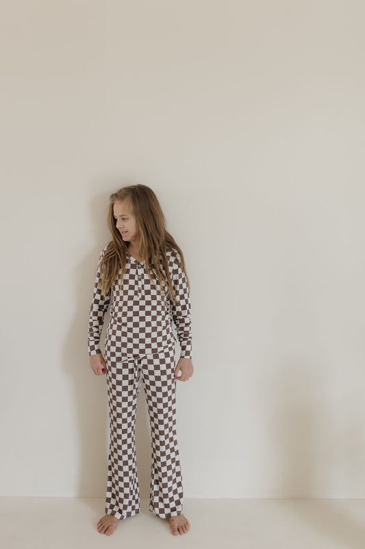 A young girl with long hair stands barefoot against a plain wall, wearing forever french baby's Youth Flare Bamboo Pajamas by Dutton. The hypoallergenic outfit, with brown and white checkered pattern, includes a long-sleeved top and pants. She gazes to the side in a relaxed posture.