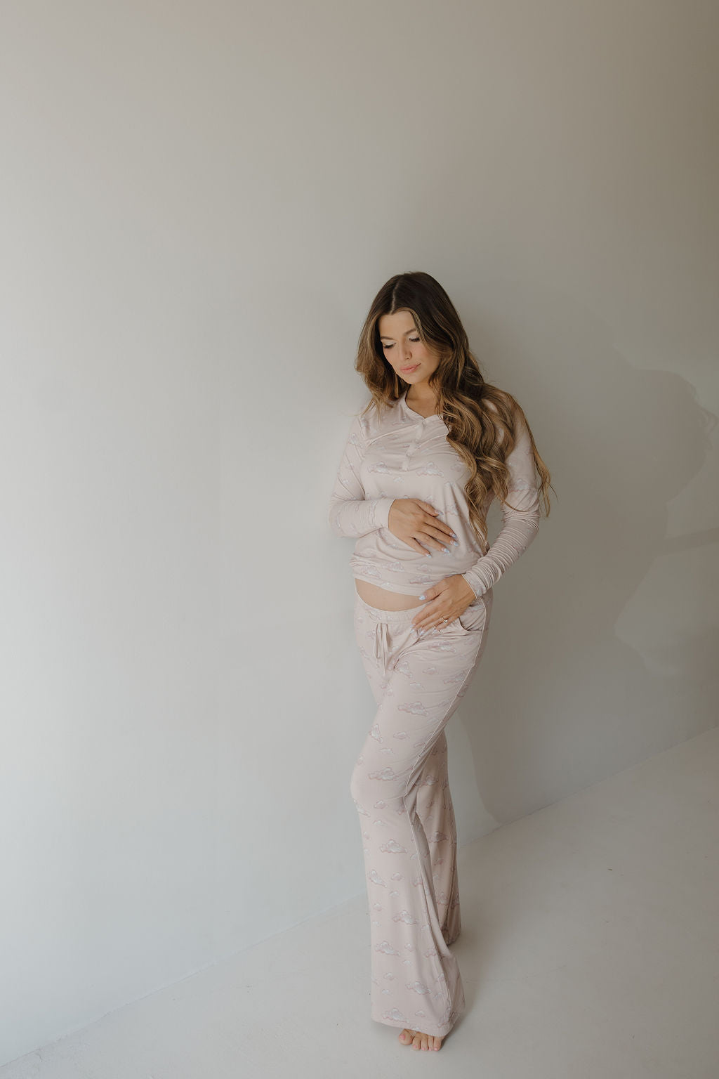 A pregnant woman with long hair stands against a light wall, gently cradling her belly. She wears the Kendy 3 Women's Bamboo Pajama | Ruby—comfortable and hypo-allergenic, ideal for breastfeeding moms seeking softness and ease.