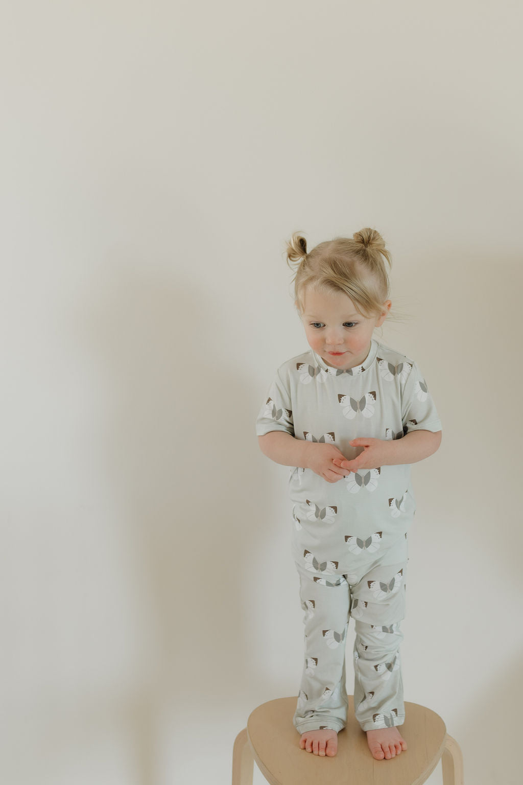 A toddler with light hair in two buns stands on a wooden stool, wearing the light gray Toddler Short Sleeve Bamboo Lounge Set with a cat pattern. Made by forever french baby from hypo-allergenic bamboo, it's set against a plain, light-colored wall.