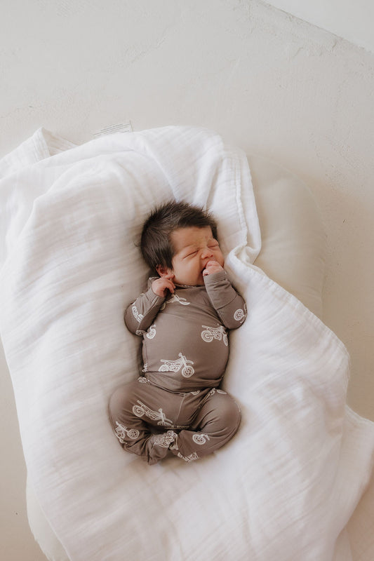 A newborn with dark hair lies on a white blanket, peacefully sucking one hand and wearing the forever french baby Sleepy Time Set | Dirt Bike hypo-allergenic brown onesie with bicycle prints.