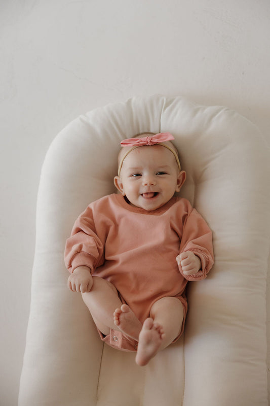 A smiling baby wearing the forever french baby Sweatshirt Romper | Rose sits on a soft, white cushion, looking up at the camera.