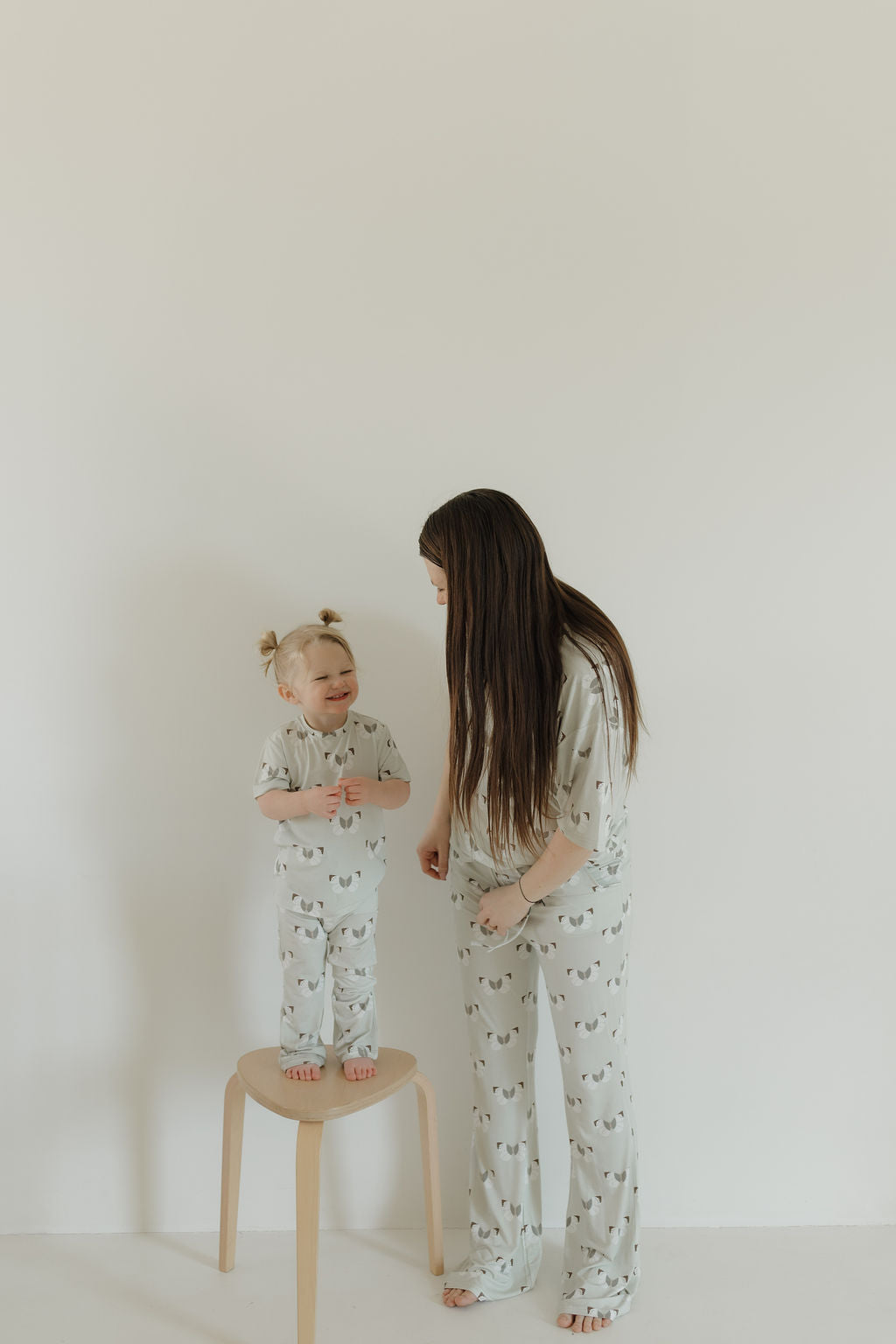 In a light, minimalistic room, a woman and toddler happily wear forever french baby’s breathable, hypoallergenic Toddler Short Sleeve Bamboo Lounge Set | Butterflies. Both stand in matching sets as the smiling toddler looks up from a wooden stool.