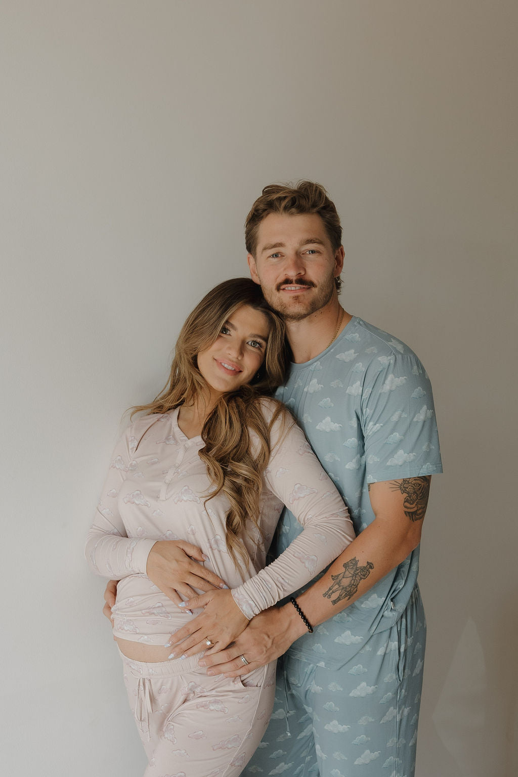 A couple poses together in matching pajamas—she wears Kendy 3’s Women's Bamboo Pajama in Ruby, ideal for breastfeeding moms, while he wears light blue. She cradles her pregnant belly and he holds her waist as they smile softly.