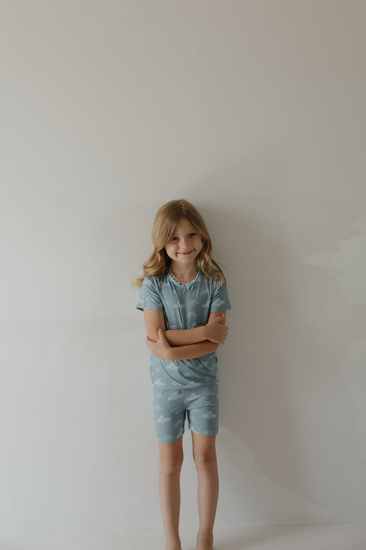 A young child smiles with arms crossed, barefoot against a plain wall, wearing the Kendy 3 Bamboo Short Set | Duke—light blue pajamas with a cloud pattern, ideal breathable sleepwear for cozy nights.