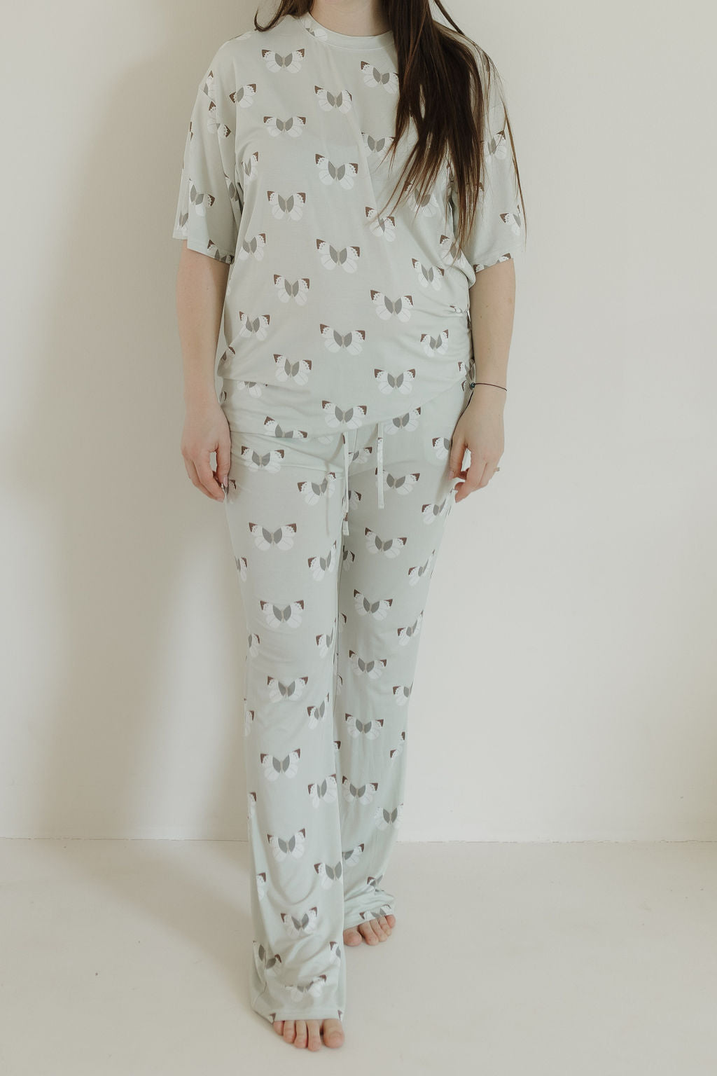 A person stands barefoot against a white background in oversized short sleeve women's bamboo pajamas by forever french baby, adorned with a playful butterflies pattern. Their long, dark hair flows down, and the hypoallergenic bamboo fabric ensures a comfortable and soothing experience.