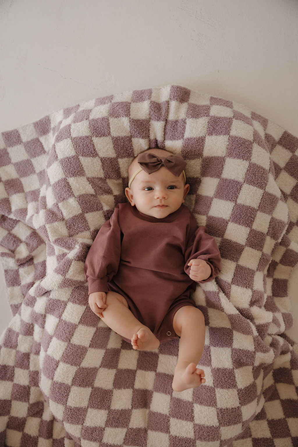 A baby in the forever french baby Sweatshirt Romper | Twilight and matching headband lies on a soft, cream and mauve checkered blanket, gazing up with a relaxed expression.