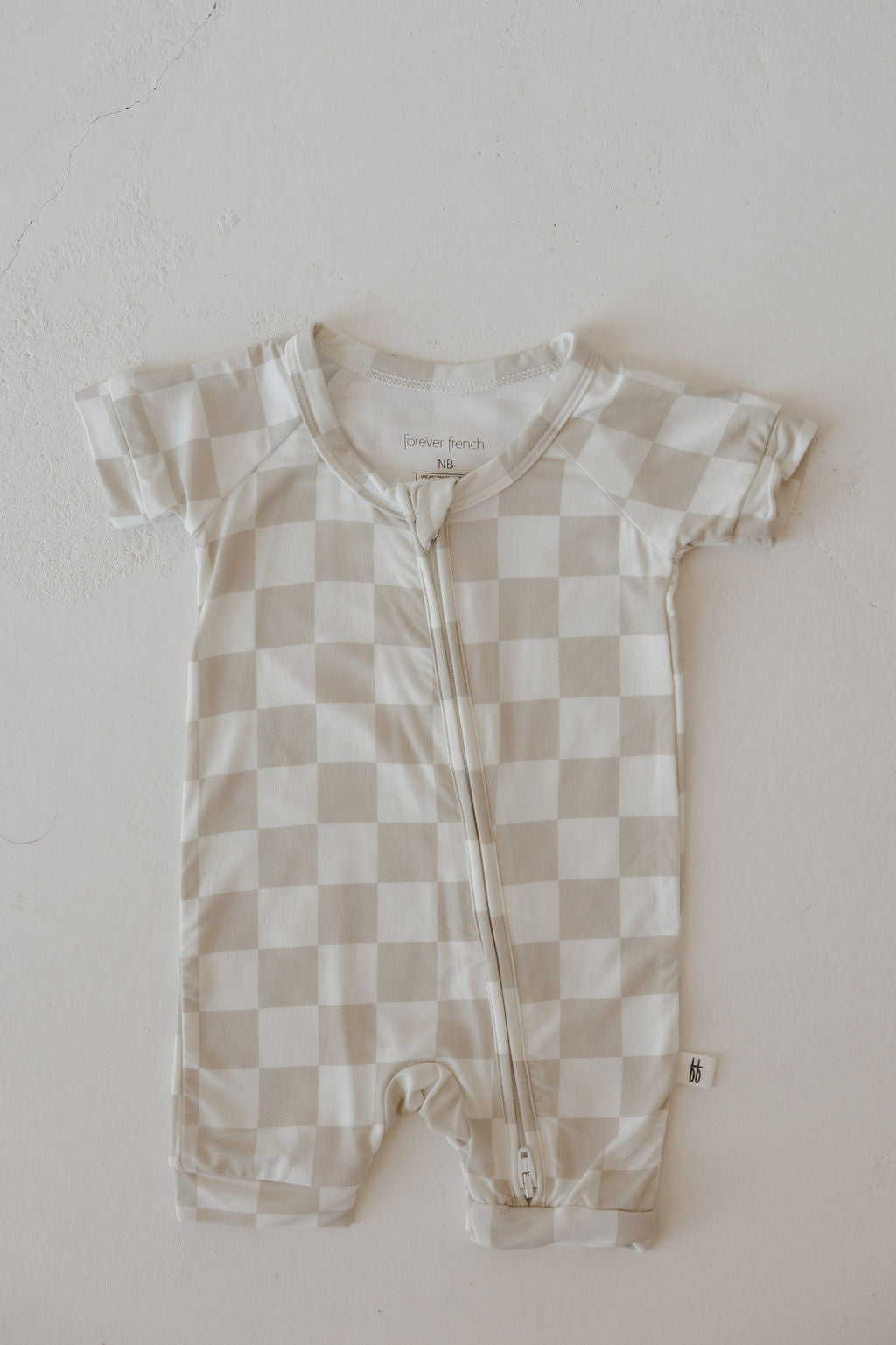 The Bamboo Zip Shortie | Checkerboard by forever french baby is a short-sleeved, beige and white checkered romper with a diagonal front zipper, made from breathable fabric and shown laid flat on a light surface.
