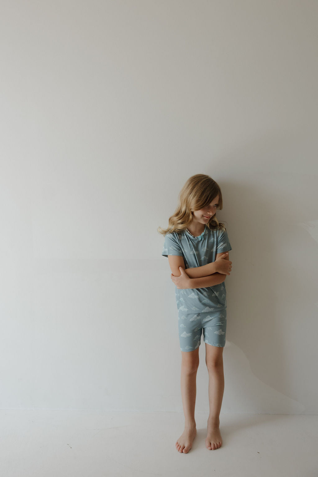 A young child with long blonde hair stands barefoot against a light wall, wearing Kendy 3’s Bamboo Short Set | Duke—breathable light blue pajamas with a subtle cloud pattern—looking down, arms crossed and expression neutral.