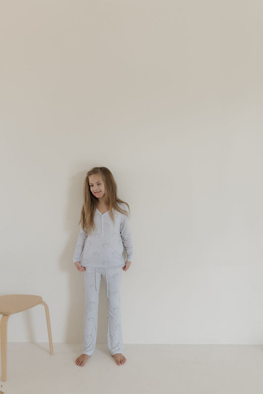 A young girl with long hair stands barefoot against a neutral wall, wearing forever french baby's Youth Flare Bamboo Pajamas | Howdy in light hues and subtle patterns. A simple wooden chair is on her left, enhancing the minimalistic and serene setting created by the hypoallergenic attire.