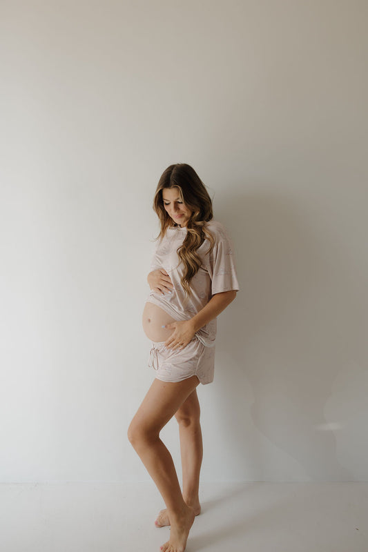 A pregnant woman with long wavy hair stands barefoot against a plain light wall, cradling her baby bump and looking down. She wears the Kendy 3 Women's Bamboo Short Set | Ruby—loose, breathable, hypo-allergenic bamboo top and shorts.