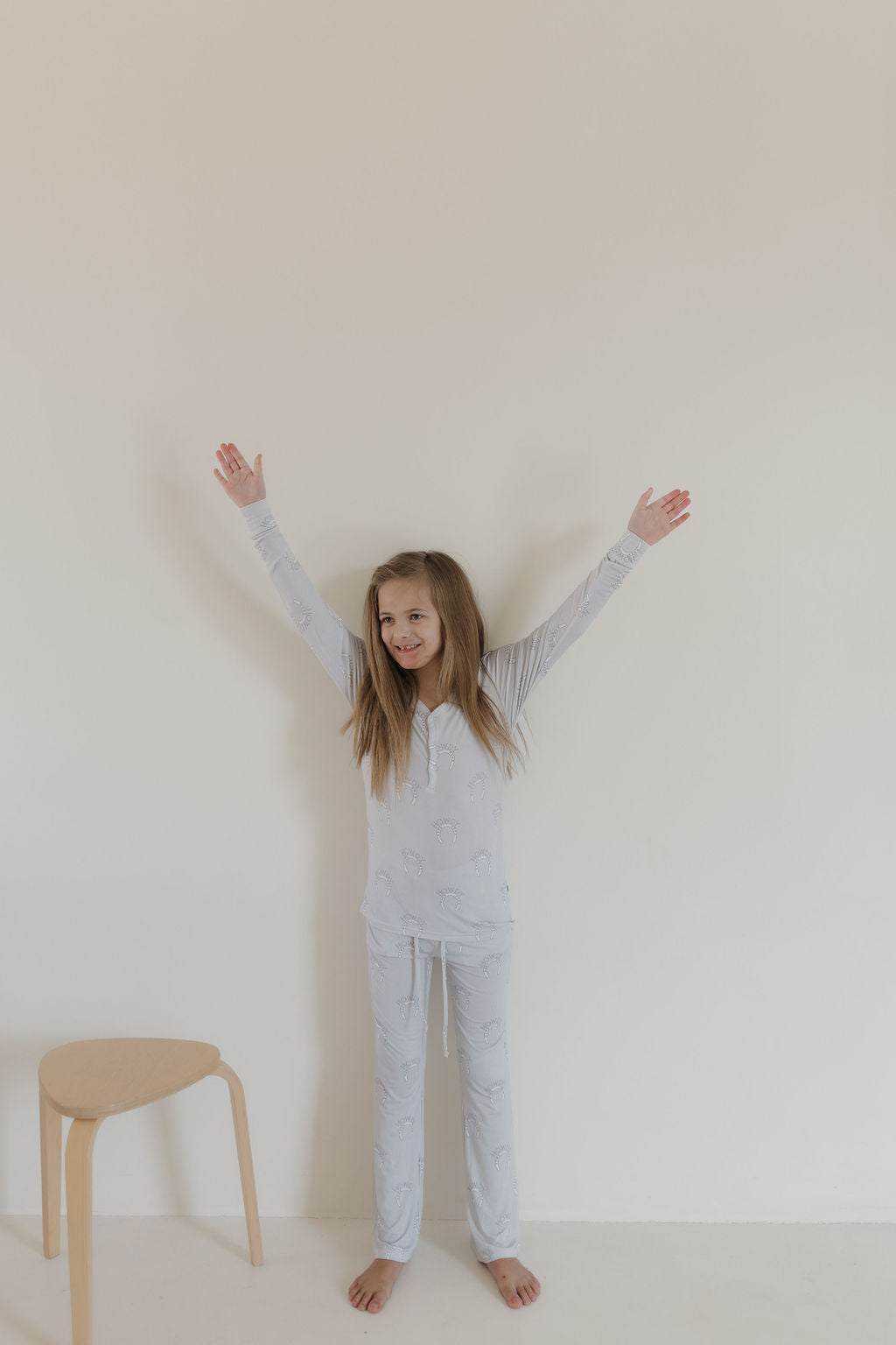 A child in light-colored Youth Flare Bamboo Pajamas by forever french baby, featuring unicorn designs, smiles with arms raised, barefoot against a plain white wall. A wooden stool stands nearby on the left.