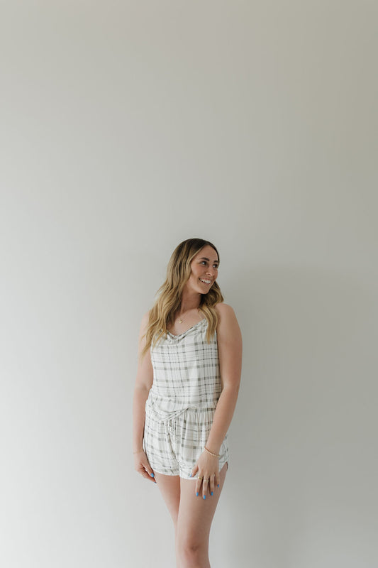 A woman smiles to her left, standing against a light backdrop in the Cami Women's Bamboo Set | Picnic Plaid by forever french baby. The sleeveless romper is made of breathable fabric. Her long hair falls in loose waves, and her arms are relaxed by her sides.