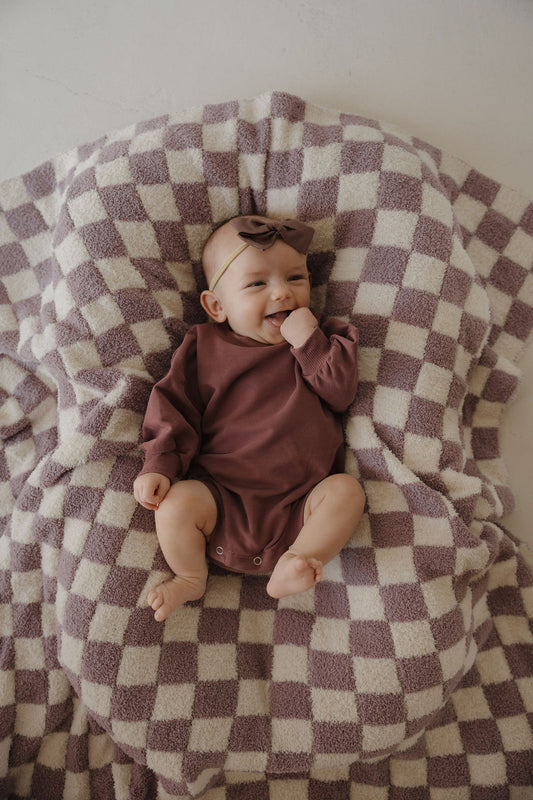 A smiling baby in the forever french baby Sweatshirt Romper | Twilight and a matching bow headband lies on a beige and mauve checkered blanket, looking happy and relaxed with one hand near their mouth.