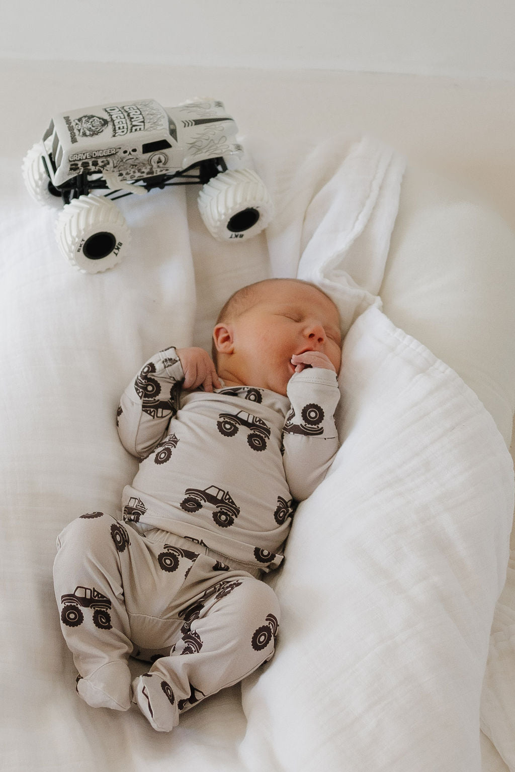 A newborn sleeps on a white blanket in hypo-allergenic bamboo pajamas with truck prints, with the forever french baby Sleepy Time Set | Monster Truck toy placed beside them.