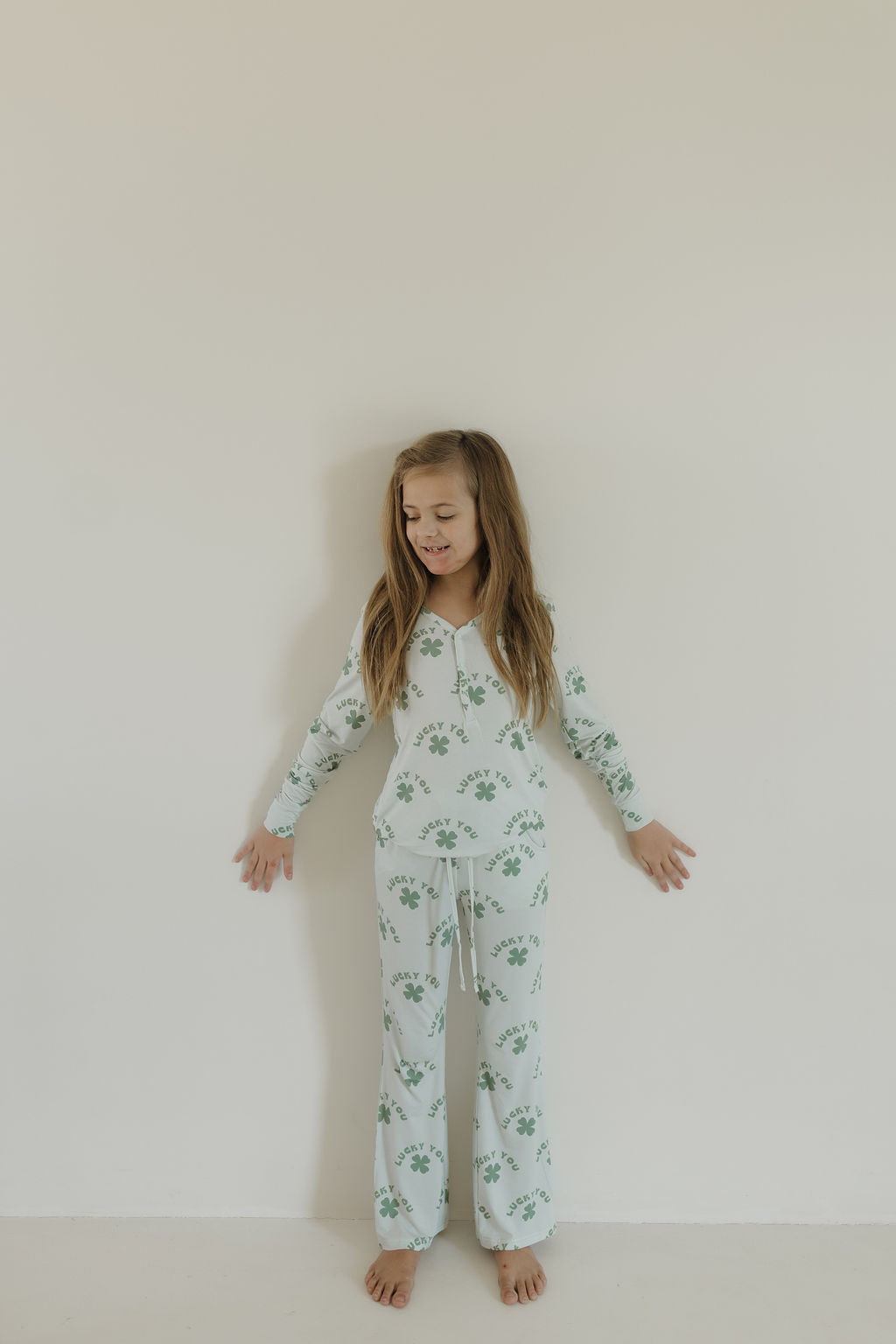 A young girl with long hair leans against a plain white wall wearing Youth Flare Bamboo Pajamas | Lucky You from forever french baby, featuring a green floral pattern. She is barefoot, her arms relaxed by her sides, and she has a calm expression.