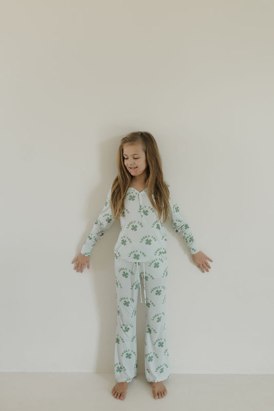 A young girl with long hair leans against a plain white wall wearing Youth Flare Bamboo Pajamas | Lucky You from forever french baby, featuring a green floral pattern. She is barefoot, her arms relaxed by her sides, and she has a calm expression.