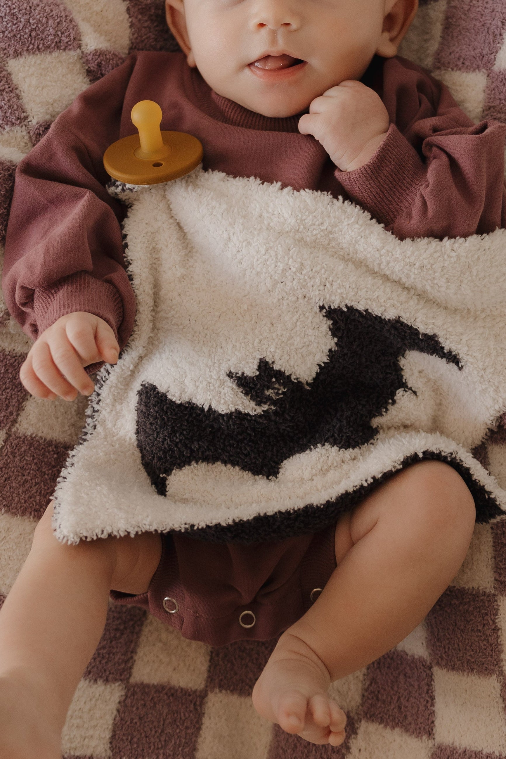 A baby dressed in maroon lies on a checkered blanket, cozily snuggled under the forever french baby Lovey | It’s Bats!—a plush microfiber white blanket adorned with a playful black bat design. A yellow pacifier rests nearby on the blanket, adding a soft touch to this forever french baby moment.