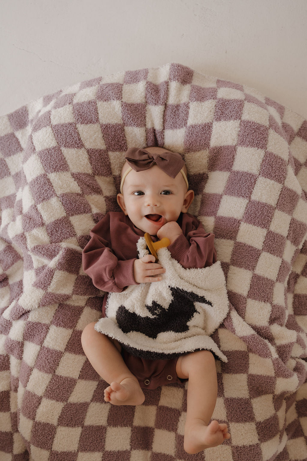 A smiling baby in forever french baby mauve clothes and headband lies on a mauve and white checkered blanket, holding the Lovey | It's Bats! plush microfiber lovey featuring a black bat design.