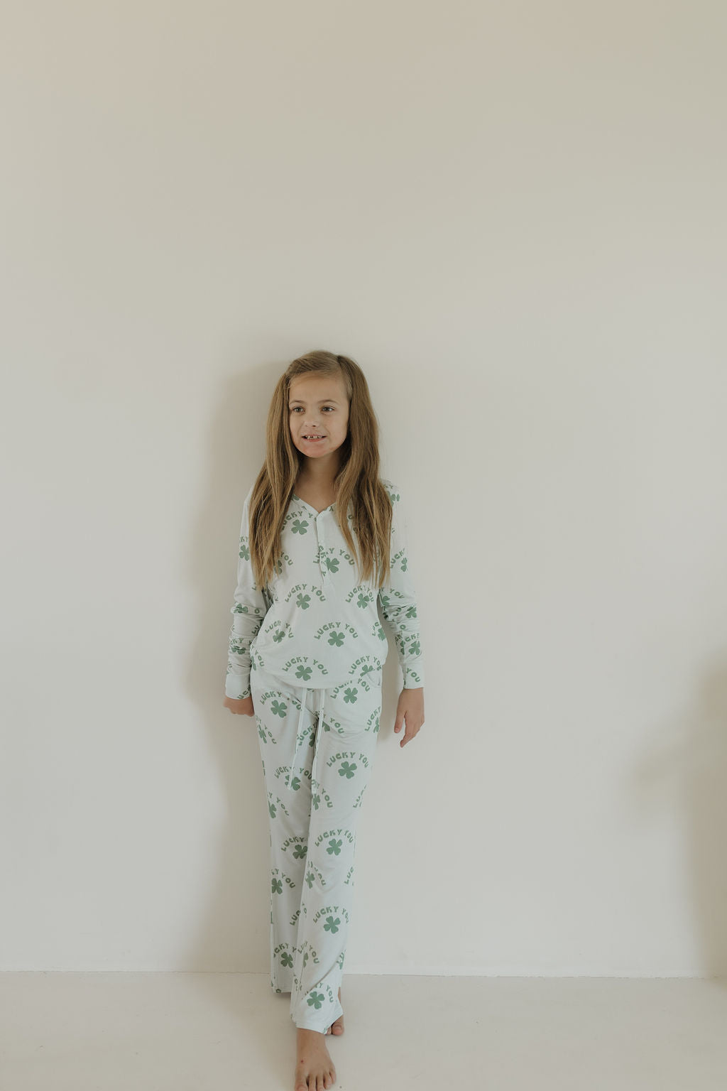 A young girl with long hair stands barefoot against a plain light-colored wall. Wearing forever french baby's Youth Flare Bamboo Pajamas in a floral print, she looks content and relaxed. The breathable, hypo-allergenic fabric ensures comfort as she enjoys tranquility.