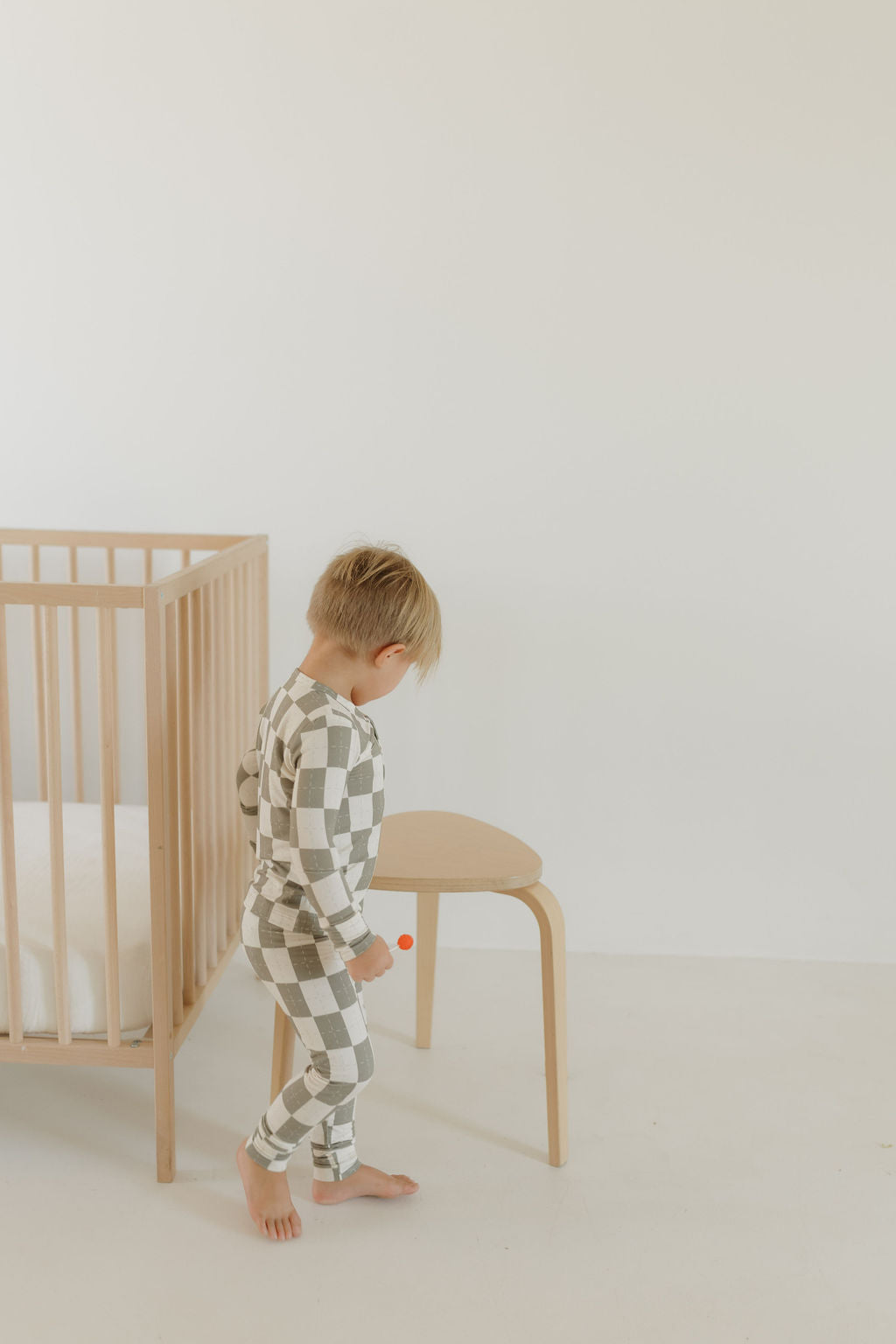 A toddler wearing "Bamboo Two Piece Pajamas | Oat & Sage" by forever french baby stands near a wooden crib and chair, holding a small orange object. The room showcases a minimalist design with light-colored walls and flooring.