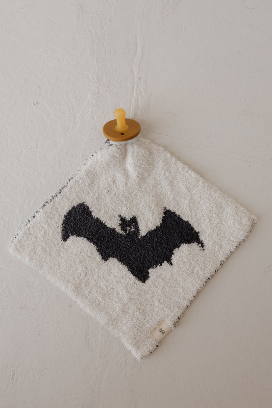 A yellow pacifier sits on a soft white cloth with a large black bat silhouette, atop plush microfiber—the Lovey | It’s Bats! by forever french baby creates the coziest scene for your little one.