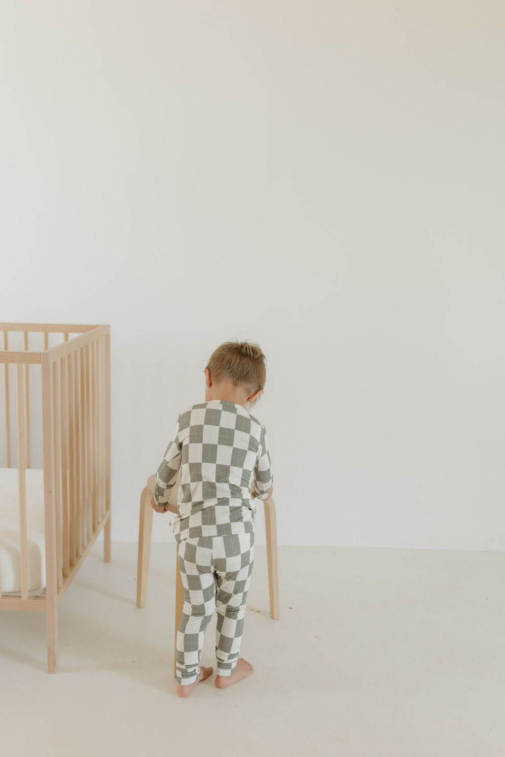 A toddler in Bamboo Two Piece Pajamas | Oat & Sage from forever french baby stands facing away, holding a wooden chair. Beside them is a light-colored crib, enhancing the room's minimalistic vibe with its plain white wall and breathable fabric accents.