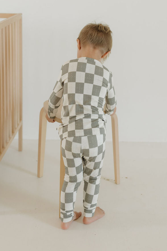 A young child in the Oat & Sage Bamboo Two Piece Pajamas by forever french baby stands barefoot, facing away and touching a light wooden stool in a minimal, light-colored room with a crib visible on the left. Crafted from breathable, hypoallergenic bamboo sleepwear fabric, these kids pajamas offer comfort and peace of mind.