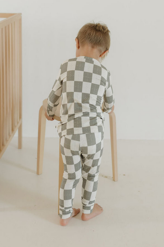 A young child in the Oat & Sage Bamboo Two Piece Pajamas by forever french baby stands barefoot, facing away and touching a light wooden stool in a minimal, light-colored room with a crib visible on the left. Crafted from breathable, hypoallergenic bamboo sleepwear fabric, these kids pajamas offer comfort and peace of mind.