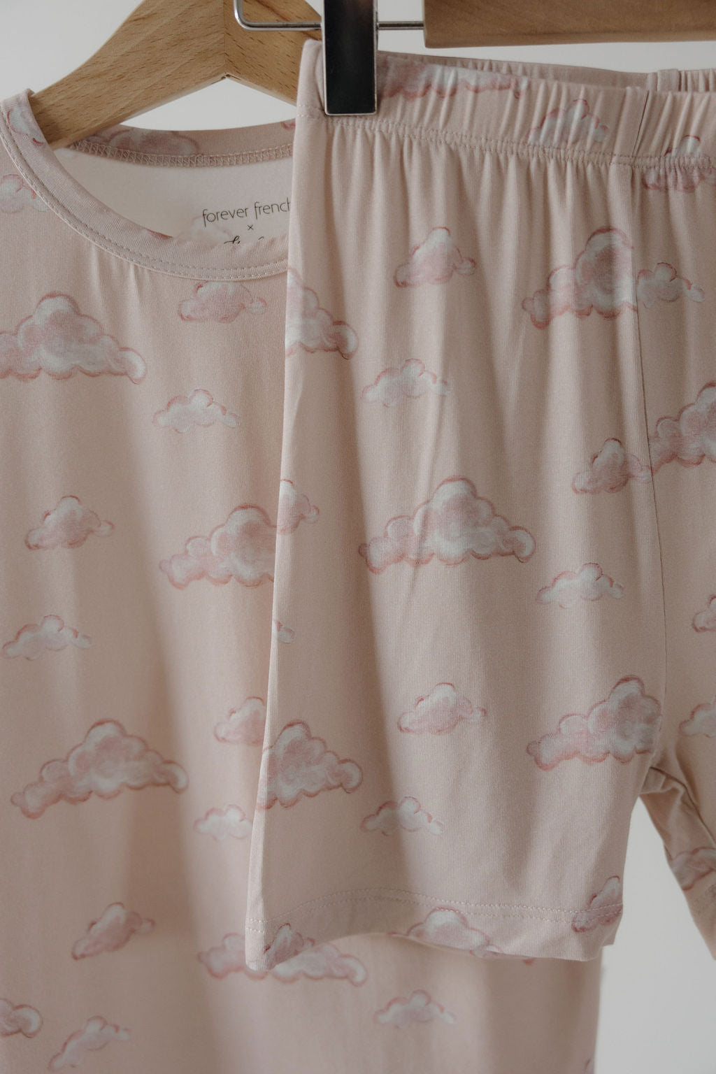 The Bamboo Short Set | Ruby by Kendy 3, featuring a short-sleeved top and elastic-waist shorts in soft pink bamboo with light pink cloud patterns, hangs on wooden hangers. The set is made from breathable fabric.