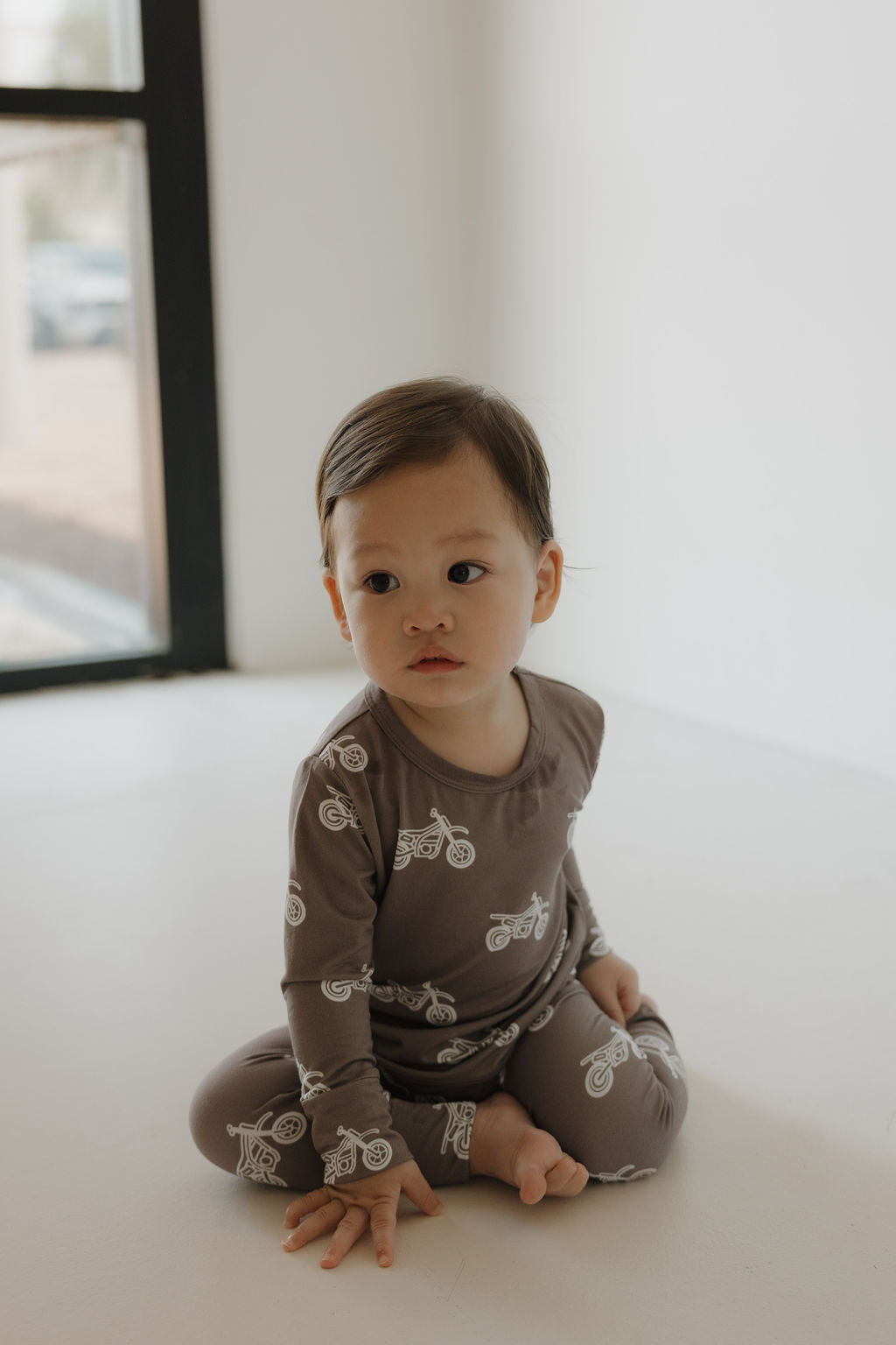A young child with short brown hair sits on the floor in a softly lit room, wearing the forever french baby Bamboo Footless Sleepy Time Set | Dirt Bike pajamas. A large window is visible in the background.