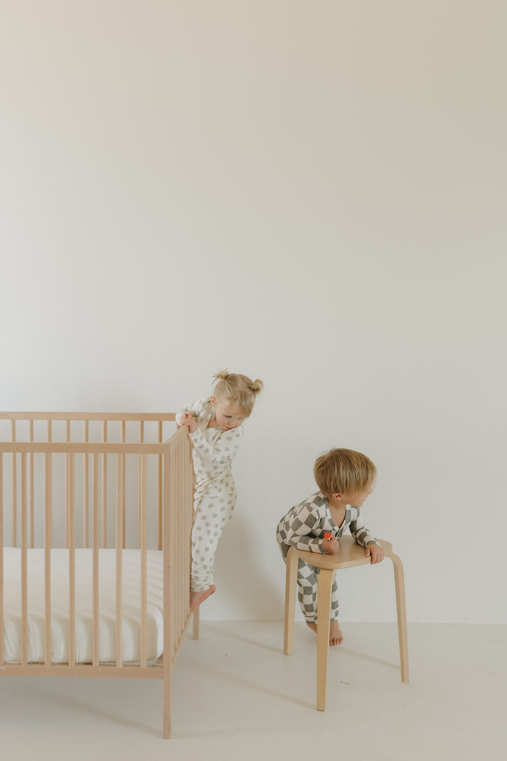 Two children in "Bamboo Two Piece Pajamas | Oat & Sage" by forever french baby are in a minimalist room. One leans on a wooden crib wearing star-patterned attire, while the other stands on a stool in checkered pajamas holding a toy.